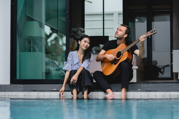 Couple lover sitting beside swimming pool romantic atmosphere playing an acoustic guitar represent a romantic getaway, a quiet weekend at home simply the joy sharing music and time with a loved one.
