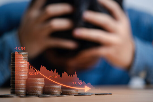 Businessman clutches his head due to stress from the great economic recession behind coins decreasing and red graph downturn concept. - Powered by Adobe