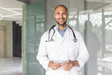 A smiling male doctor in a white coat, holding a tablet, standing in a modern healthcare setting....