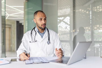 A doctor looks at his laptop while consulting medical documents in an office setting, indoors. He...