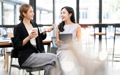 Two asian business women sit at desk discuss project details, diverse female colleagues met in office, share opinion