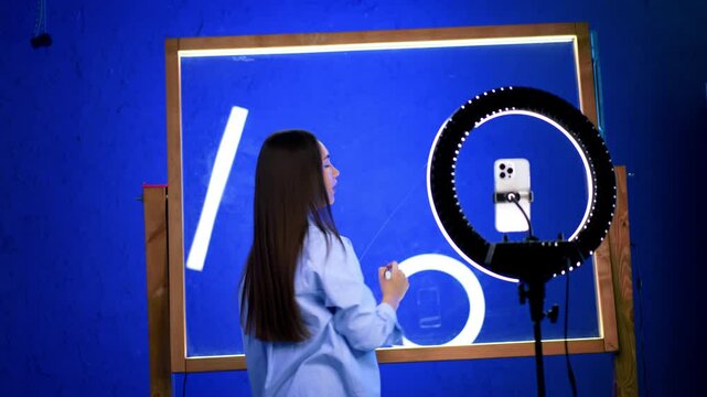 Creative woman filming art. A young woman creates art while filming with a smartphone in a brightly lit blue studio space.