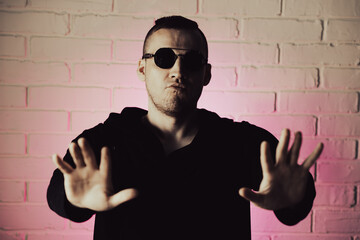Stylish man poses confidently against a pink-lit brick wall while wearing sunglasses and a dark...