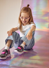 A cheerful young girl with long hair tied into a small bun sits on a colorful, iridescent background and laughs. She is wearing pink glasses, a white T-shirt and light blue jeans.