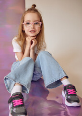 A charming young girl with long hair tied in a bun and glasses sits with her chin resting on her hands against a colorful gradient background. She is wearing wide jeans and black sneakers.