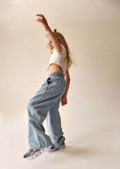 An energetic young girl stands in a dynamic pose, raising one arm up. Her long blonde hair flies to the sides. The child in a white crop top and jeans tries to keep her balance, standing on one leg.