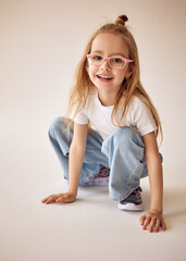 An active and cheerful young girl with long blond hair and glasses sits on her haunches and smiles. She touches the floor with her hands. A child in a white T-shirt and blue jeans poses on a cyclorama