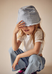 A cute seven-year-old child in a light blue headdress that partially covers her eyes is squatting. The little girl in a white T-shirt and jeans playfully touches the brim of her hat with her hand.