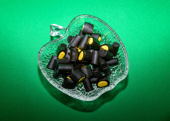 A glass bowl filled with black licorice candies featuring vibrant yellow centers is displayed against a vivid green background. These treats bring a playful touch to the gathering