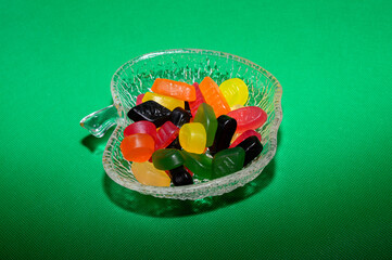 A clear decorative bowl holds a variety of colorful jelly candies, including orange, yellow, green, and red. The vibrant colors contrast against a bright green backdrop