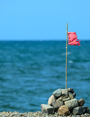 A tattered red flag is perched on a stick, resting atop a pile of stones near calm blue waters. The clear sky enhances the serene coastal atmosphere during daylight