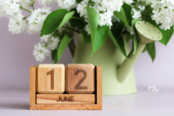 A wooden calendar showing June 12 and a white lilacs in a decorative watering can. the date of an...