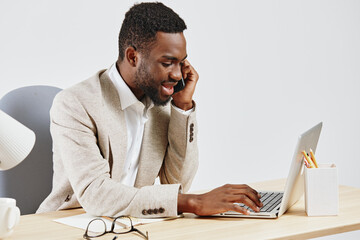 Smiling young man working on a laptop at a desk in a modern office setting, wearing a beige cardigan and white shirt, with glasses and stationery nearby, conveying productivity and professionalism
