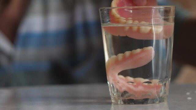 an elderly person's hand takes removable dentures out of a glass and puts them on, removable dentures in a glass with water, tooth loss and prosthetics