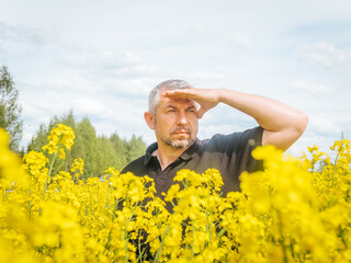 A middle-aged man shields his eyes from the sun while standing in a vibrant yellow rapeseed field under a bright sky with scattered clouds.