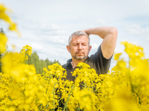  A man with a contemplative expression stands in a blooming rapeseed field, one hand resting behind his head, under a soft, partly cloudy sky.