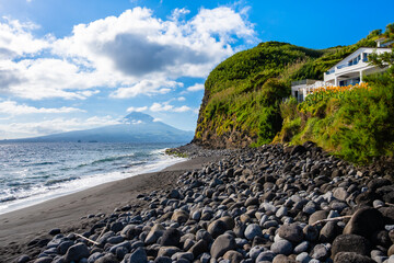 Stones on Almoxarife black sand volcanic beach with blue ocean and houses on cliff on coast of Faial island, Azores, Portugal