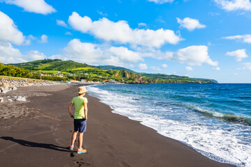 Woman tourist standing on black sandy volacnic Almoxarife beach with azure ocean water on coast of Faial island, Azores, Portugal