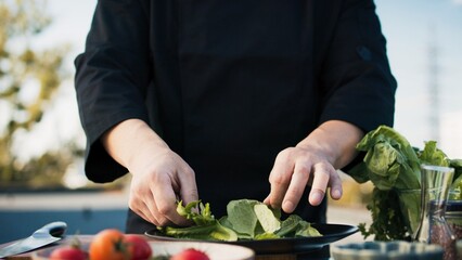 Asian chef adding ingredients to plate and seasoning outdoor. Man in uniform garnishing salad on...