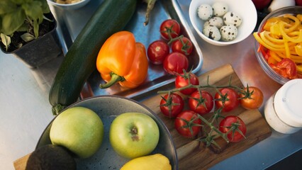 Fresh vegetables and fruits arranged on kitchen table outdoors. Colorful ingredients including tomatoes, zucchini, apples, and bell peppers, quail eggs. Close-up of vibrant food setup for cooking.