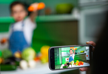 Fototapeta na wymiar Young chef captures cooking moment. A child holds a vegetable while posing for a video in a bright kitchen filled with fresh produce.
