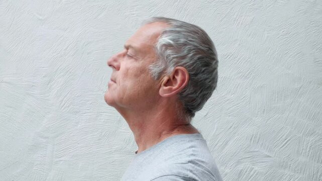 Left facing profile of a middle aged white man doing ten repetitions of back and fore cervical vestibular neck physiotherapy exercises against a textured wall.