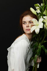 A serene woman of Eastern European descent holding white lilies, wearing a stylish white blouse,...