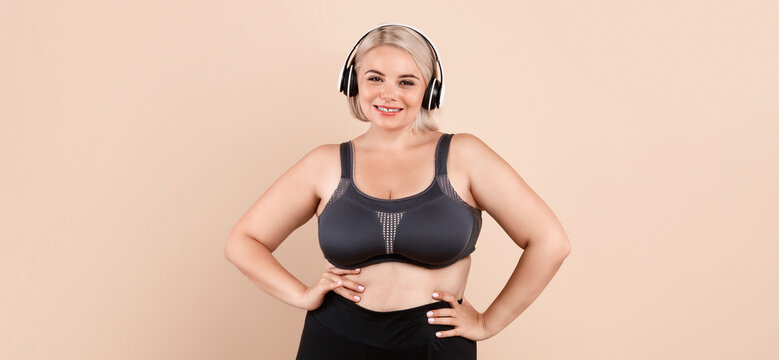 Fototapeta Weightloss Concept. Happy Plus Size Woman In Sporswear Posing Over Beige Studio Background, Curvy Young Lady Wearing Wireless Headphones Ready For Training, Smiling At Camera, Copy Space