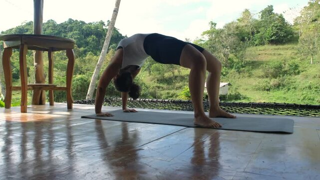 In a serene setting, a young woman performs a bridge pose on her yoga mat, enjoying the tranquil beauty of lush rice paddies and vibrant tropical nature on a sunlit terrace, slow motion