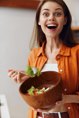 Happy young woman enjoying a healthy salad in a wooden bowl, wearing a bright orange shirt, set in a modern kitchen with a warm color palette
