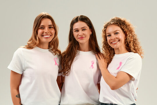 Three women showing support for breast cancer awareness wearing white shirts with pink ribbons
