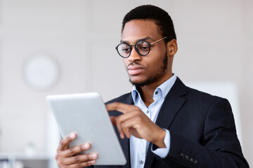 Plakat Closeup of serious black entrepreneur in glasses using modern digital tablet, checking trades and stocks online, office interior, copy space. Young african american manager holding digital pad