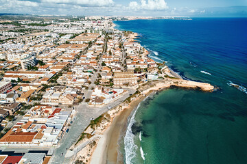 High-angle drone photo of Spanish seaside town with residential areas, clear turquoise water, sandy beaches, and rocky shoreline. Torre de la Horadada on Costa Blanca, Spain