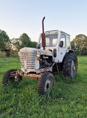 An old white tractor on the green grass in the countryside. A rusty agricultural tractor with a cab stands on the grass among the trees. A symbol of rural street machinery and manual labor.