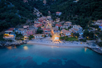 Aerial view of a coastal village illuminated at twilight, nestled by the sea and lush greenery.