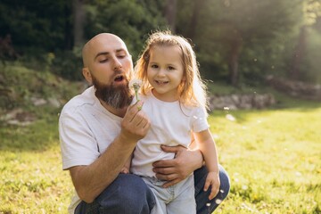 Father and daughter blowing dandelion in a park during a sunny day