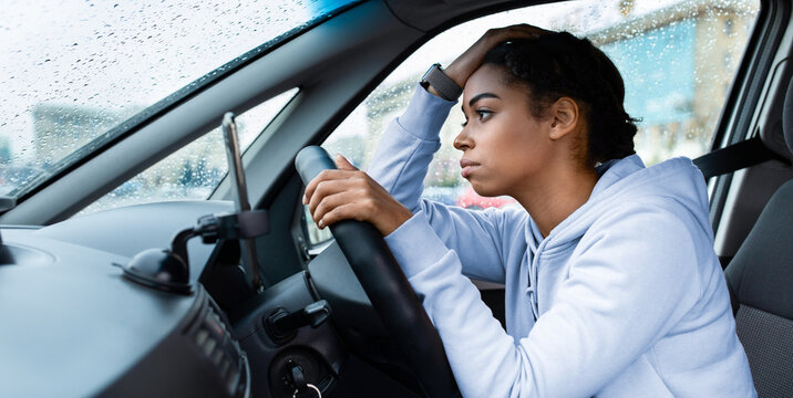 Rainy weather, problems on road and traffic jams. Sad tired millennial african american lady with smartwatch at steering strapped up, with hand on head, looks at wet window, side view, free space
