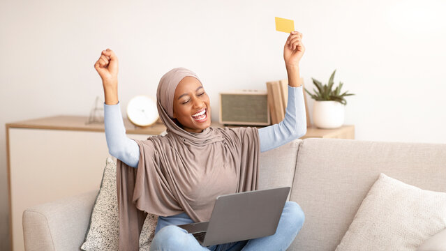 African American woman in hijab raising her hands up in excitement over huge discount in online shop, buying goods over internet on sale, using laptop and credit card a home