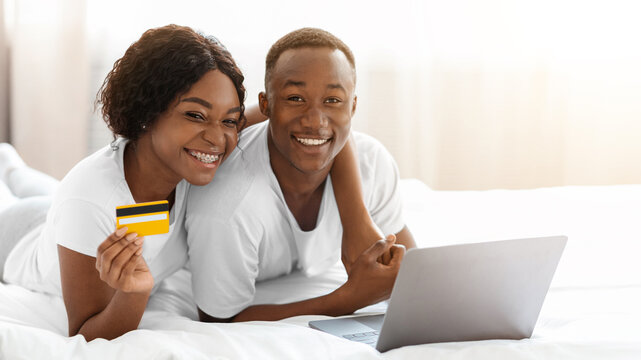 Young black couple laying on bed with laptop, shopping online, recommending credit card, closeup, copy space. Smiling african american man and woman in pajamas making online order, using computer
