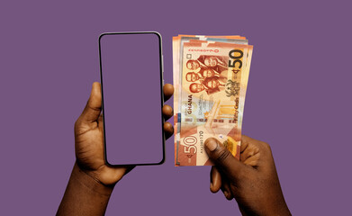 Black African hand holding mobile smart phone with blank screen and Ghanaian cedi notes in another hand over purple background 
