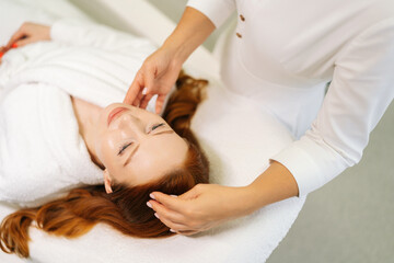 Fototapeta premium Top view of beautician gently touching female client face and hair while woman lying relaxing on massage table, preparing for rejuvenating facial treatment in serene spa environment.
