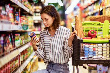 Consumption And Consumerism. Portrait Of Young Woman With Shopping Cart In Market Buying Groceries Food Taking Products From Shelves In Store, Holding Glass Jar Of Sauce, Checking Label Or Expiry Date