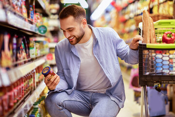 Consumption And Consumerism. Portrait Of Smiling Bearded Man With Shopping Cart In Market Buying Groceries Food Taking Products From Shelves In Store, Holding Glass Jar Of Sauce, Checking Label