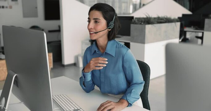 Young Latin customer support service employee talking on video call, using wireless headset with mic and desktop monitor in office, giving online consultation
