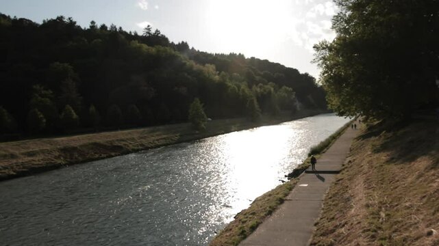 The Savinja River in Celje glows in the evening light. People walk along the riverside path, surrounded by forest and calm nature. View toward alec from the riverbank at sunset.