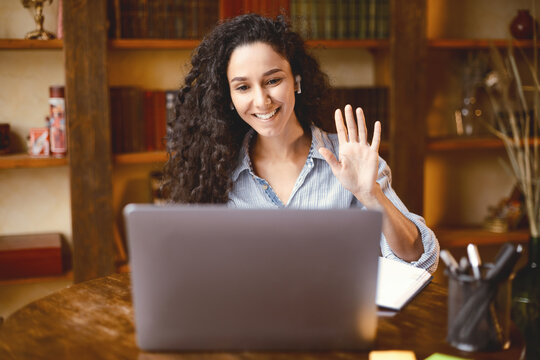 Fototapeta Teleconference And Telecommuting. Portrait of smiling woman sitting at desk, having video call on laptop, waving to webcam. Distance Education. Cheerful lady wearing headphones at virtual meeting