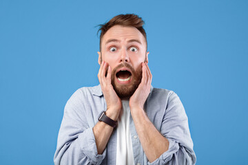 Scared young guy screaming in panic over blue studio background. Horrified bearded man feeling afraid, suffering from panic attack, yelling in fear, being dramatic