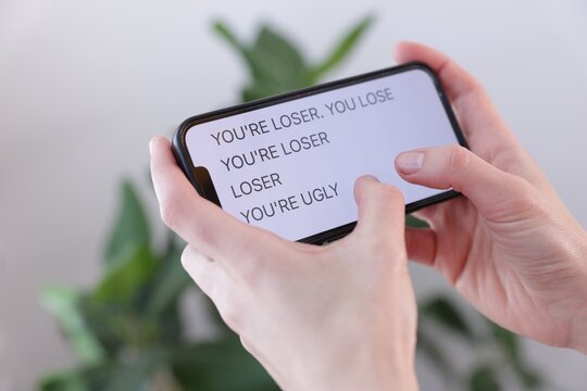 Cyber bullying. Woman holding smartphone with insults on blurred background, closeup