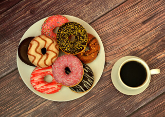 Cup of black coffee on a saucer and a plate with an assortment of fresh donuts in colorful glaze on a wooden table.