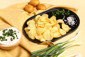 Bowl of tasty sour cream with sliced green onion and potato chips on yellow background
